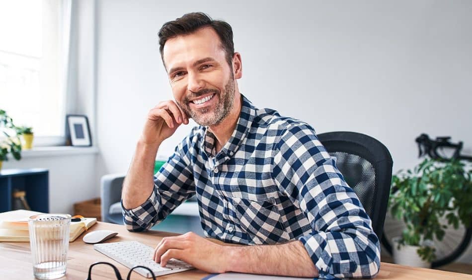 A man in a black and white checked button-up sitting at a desk and smiling