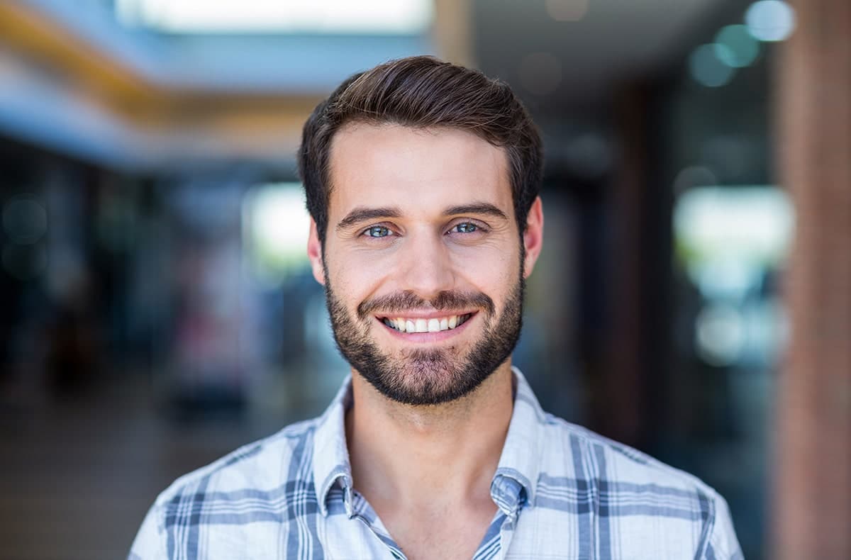 An adult man with short hair and a beard wearing a plaid button-up shirt and smiling