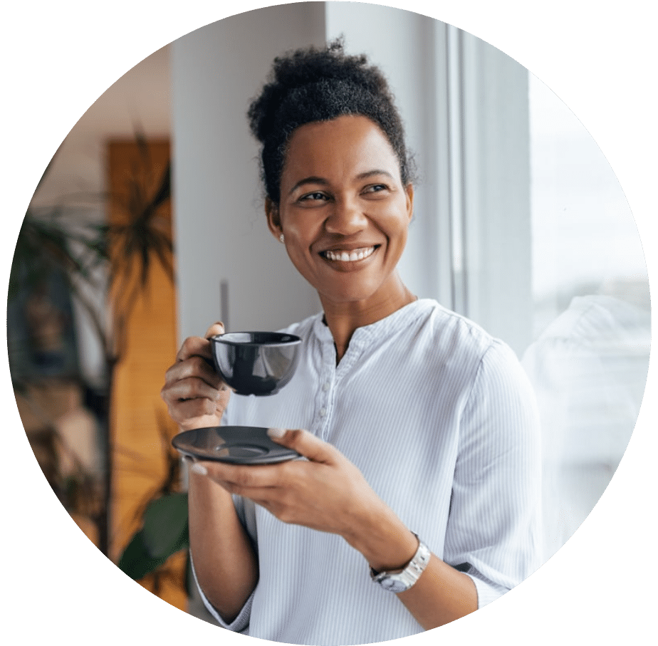 A middle-aged woman in a white button-up sipping tea next to a window smiling